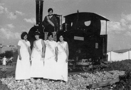 Reina y damas de fiestas junto a una locomotora en Ojos Negros.